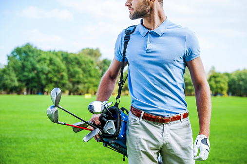 Cropped image of male golfer carrying golf bag with drivers while walking by green grass