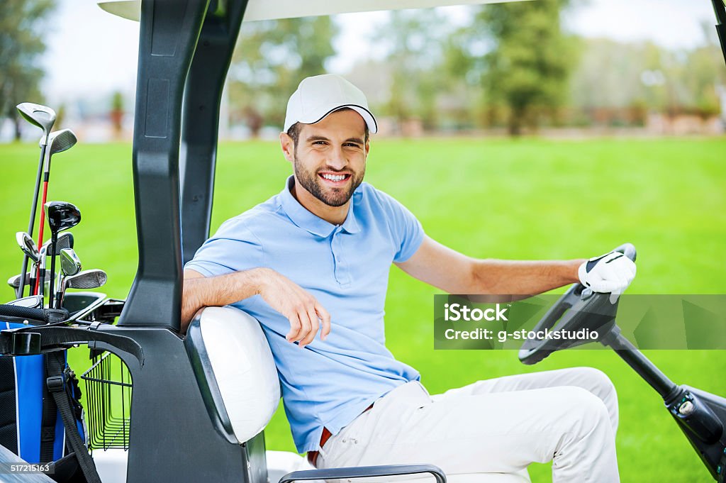 On my way to the next hole. Side view of young happy male golfer driving a golf cart and looking at camera Driving Stock Photo