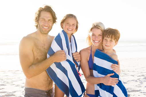 Family with their children draped in towel at beach after swim. Portrait of smiling parents with daughter and son after swimming. Family with two children looking at camera.