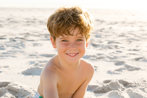 Portrait of young boy sitting on sand at beach and looking at camera. Little boy enjoying holiday at beach. Cute child playing at beach at sunset.