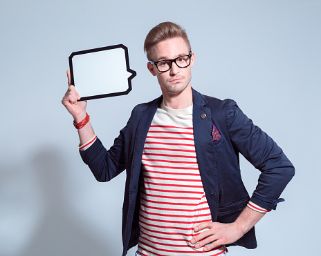 Portrait of surprised blonde young man wearing navy blue jacket, striped blouse and nerd glasses, holding a speech bubble in hand, looking at camera.