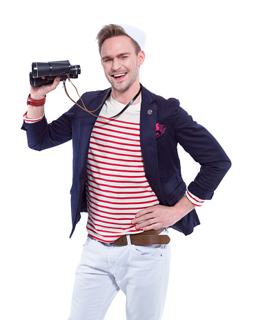 Summer portrait of an attractive blonde young man in sailor style outfit, holding binoculars in hand and laughing at camera. Studio shot, white background.
