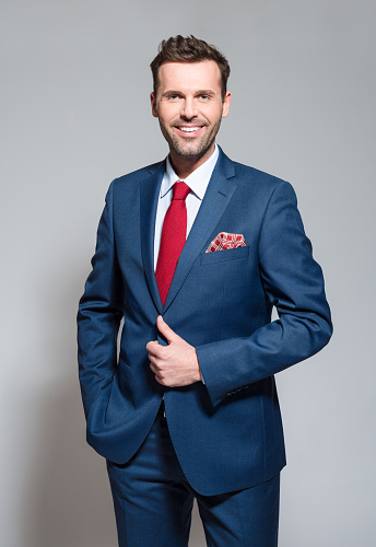Elegant businessman wearing suit, red tie and pocket square, standing against grey background and smiling at camera. Studio shot.