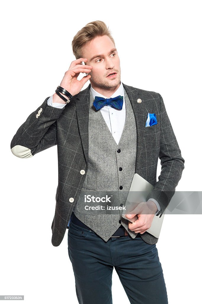 Fashionable young businessman in classical outfit talking on phone Portrait of fashionable, elegant young businessman wearing tweed jacket and bow tie, holding a laptop in hand and talking on phone. Studio shot, one person, isolated on white. Adult Stock Photo