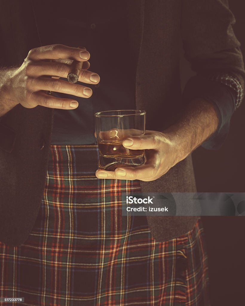 Man drinking whiskey and smoking cigar, close up of hands Elegant man wearing tweed jacket and tartan skirt, holding glass of scotch whisky and cigar. Close up of hands, unrecognizable person. Dark tone, black background. Plaid Stock Photo