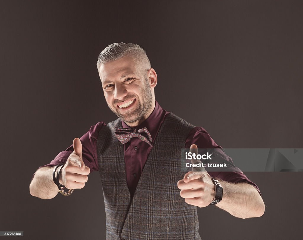 Successful elegant man wearing bow tie and tweed vest Happy elegant bearded man wearing tweed vest, shirt and bow tie, smiling at camera with thumbs up. Dark tone, black background. Adult Stock Photo