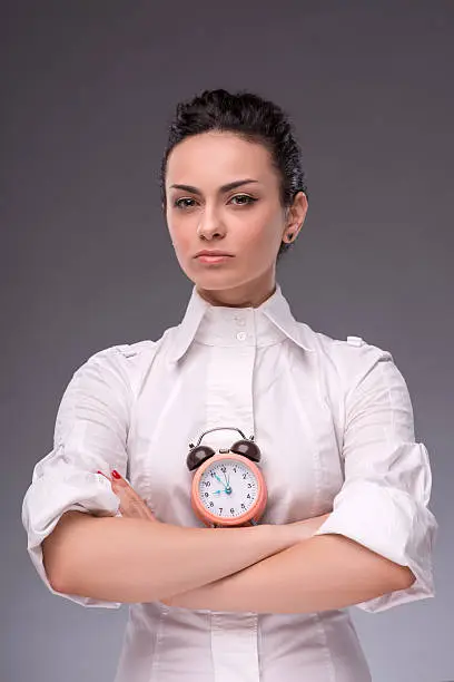 Close-up portrait of pretty girl with surprised face holding an alarm clock on her head and looking at the camera with a fright, with copy place isolated on grey background, concept of time management