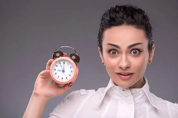 Close-up portrait of pretty girl with surprised face holding an alarm clock in her hand looking at the camera with copy place isolated on grey background, concept of time management