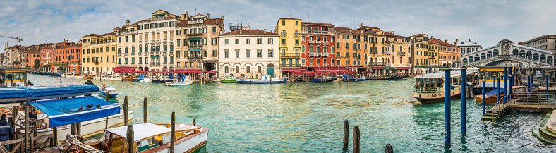 Venice, Italy - 11th March 2013: Locals and tourists on the iconic stone arch of Rialto Bridge above the broad sweep of the Grand Canal between the busy restaurants, historic palazzo, gondolas water busues and boats of the San Marco and San Polo quarters of Venice, Italy. Composite panoramic image created from ten contemporaneous sequential photographs.