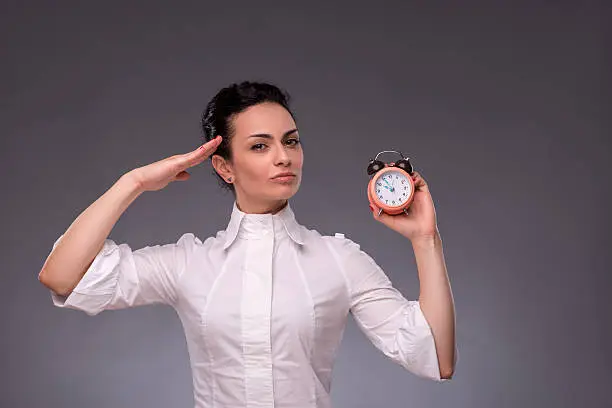 Waist-up portrait of beautiful girl looking at you while holding an alarm clock in her hand and saluting with her other hand, with copy place isolated on grey background