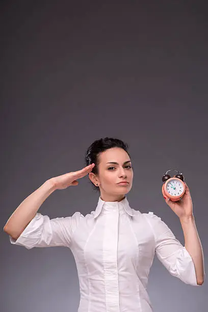 Waist-up portrait of beautiful girl looking at you while holding an alarm clock in her hand and saluting with her other hand, with copy place isolated on grey background