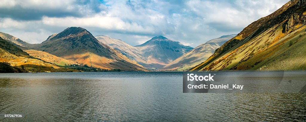 Mountain Range At Wastwater Lake With Dramatic Clouds. A wide panoramic stitch of the beautiful view, voted 'England's favourite view' at Wastwater Lake in the Lake District, UK. The image features dramatic clouds with sun shining through and illuminating parts of the mountain range. Backgrounds Stock Photo