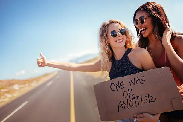 Caucasian Hipster girl and mixed race hipster girl in the daytime stand on side of two lane highway hitchhiking while holding cardboard sign with the One Way or Another written on it and sticking out thumb