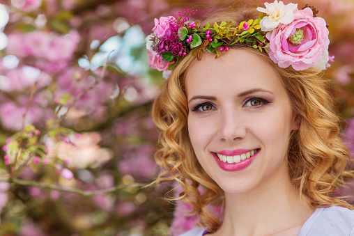 Smiling woman with blond hair with flower wreath against pink tree in blossoom, spring nature