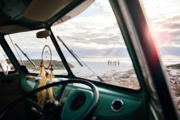 An interior window shot from a retro van window perspective looking out onto a sandy beach where, from a distance, a group of friends are walking towards a sun ray lit ocean