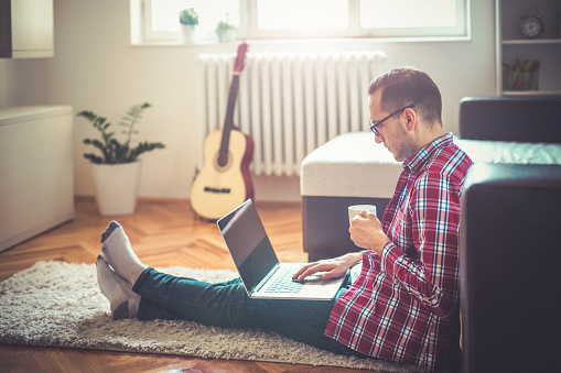 Young man sitting on the floor drinking coffee and surf the internet with lap top.