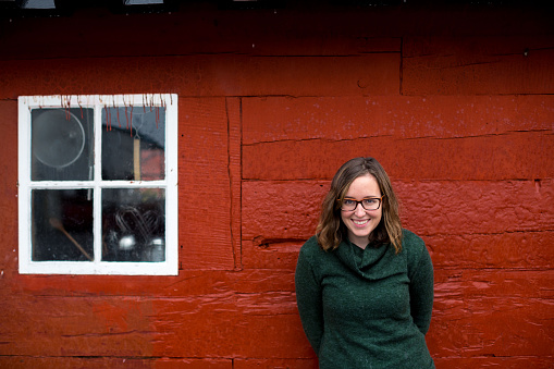 Woman, wearing a green sweater, blue hat and glasses poses in front of a classic Icelandic hut in the northern part of the country.
