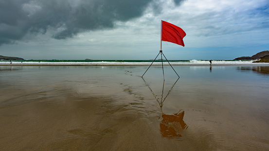 A beach in northern Europe, with a big red flag reflected on the wet sand, warning bathers against rough conditions