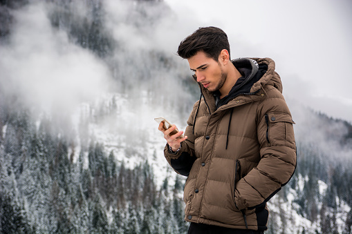 Dark haired handsome young man in winter outerwear using cell phone or smartphone, outdoor at mountain with snowy landscape behind