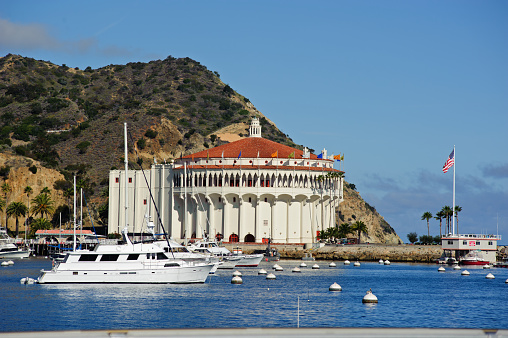 This is an image of the Casino in Avalon Harbor, Catalina Island taken from the opposite side of the harbor. There are private boats in the foreground and a hill in the background. 