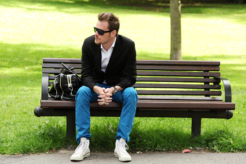 Attractive man in blue jeans and black blazer sitting on a bench in a park.