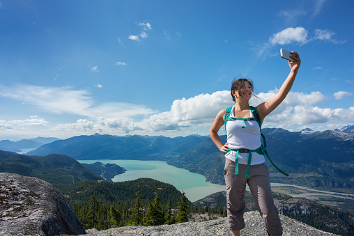 A young, female Asian hiker stands on a mountaintop and takes a selfie with her smartphone.  Squamish, British Columbia, Canada.  View of the valley, water and surrounding mountains in the background.