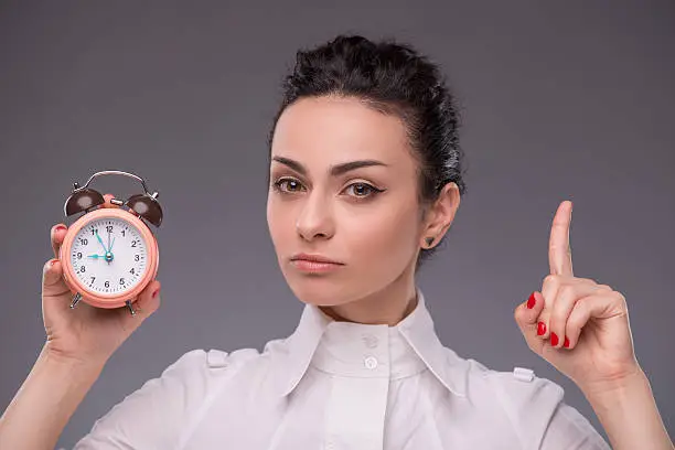 Close-up portrait of pretty girl with concentrated face holding an alarm clock in her hand showing sign Attention looking at the camera with copy place isolated on grey background