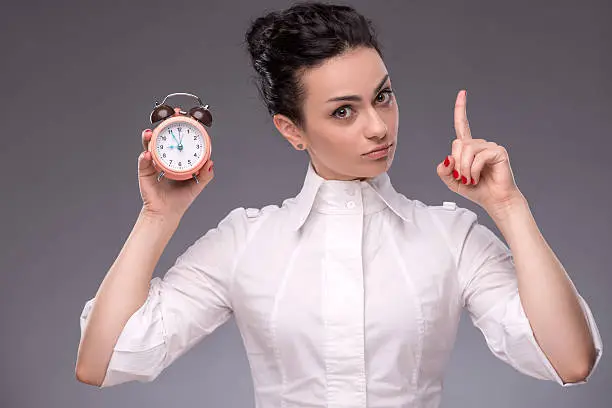 Close-up portrait of pretty girl with concentrated face holding an alarm clock in her hand showing sign Attention looking at the camera with copy place isolated on grey background