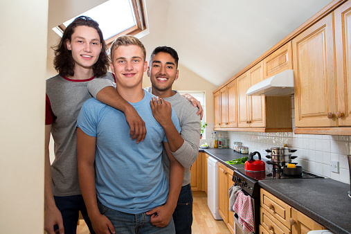 Group of male students embrace each other while standing in the kitchen and smile at the camera