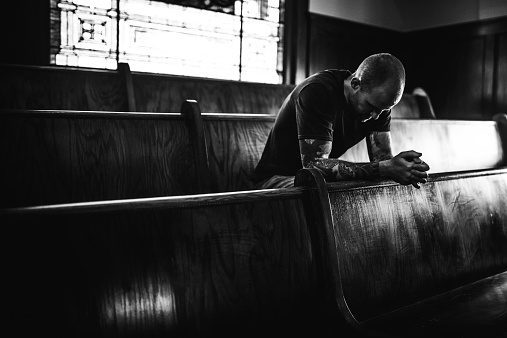 A black and white image of a man bowing his head in prayer while sitting on a wooden pew in a traditional church building. Horizontal image with copy space.