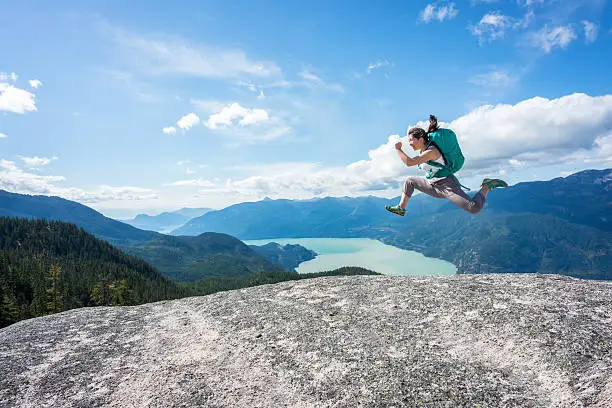 An athletic, female, Asian hiker leaps on mountain summit.  Squamish, British Columbia, Canada.  View of the valley, water and surrounding mountains in the background.