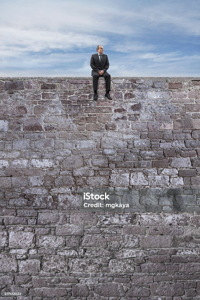 Businessman Sitting on Wall with Legs Hanging Businessman sitting on the top of a high stone wall with legs hanging over side. Adult Stock Photo