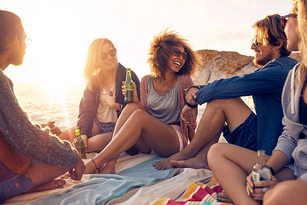 Group of smiling friends chilling on the beach Mixed race friends having fun at the beach. Group of happy young people sitting together at the beach talking and drinking beers. beach party stock pictures, royalty-free photos & images