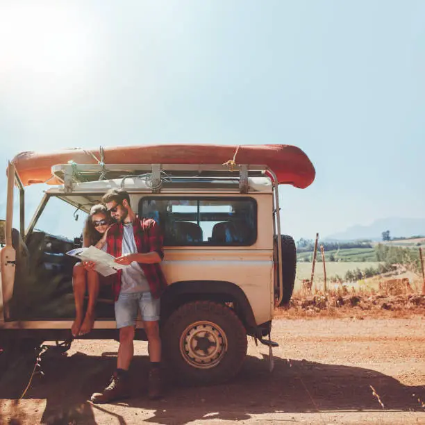 Young couple stopping on country road and looking at the map for directions. Man and woman on a road trip in countryside on a summer day.