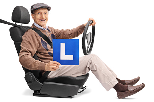 Cheerful senior holding an L-sign and holding a steering wheel seated on a car seat isolated on white background