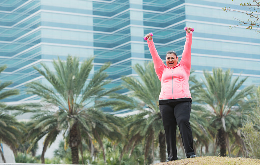 An Hispanic overweight woman exercising with handweights outdoors. She is excited and happy to have reached a goal, arms raised in the air to celebrate her success.