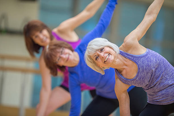 Stretching at the Gym Three senior adult women are taking a yoga class together at the gym. One woman is smiling and looking at the camera. long sleeved recreational pursuit horizontal looking at camera stock pictures, royalty-free photos & images
