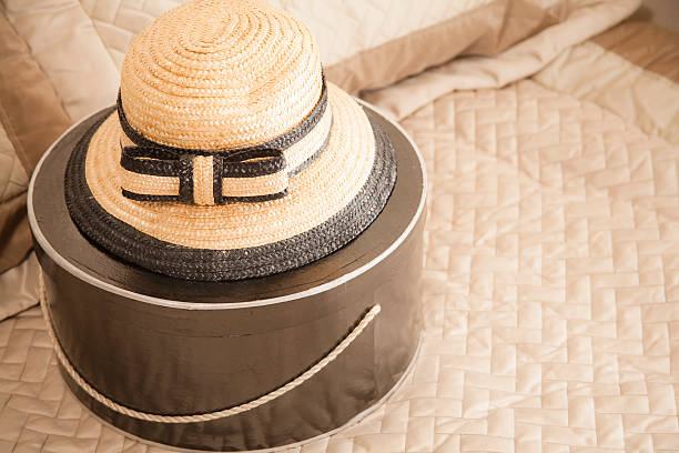 Woman's straw hat in tan and black colors and a black, round hat box lie on top of a bed in a bedroom setting.  No people.  Copyspace.  Retail, shopping, consumerism, tourism, travel, vacation concepts.