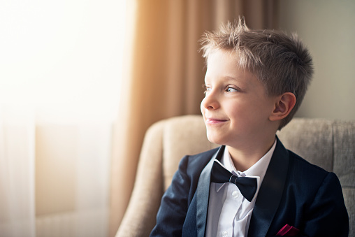 Portrait of a smiling cute blonde boy wearing a tuxedo and bow tie. The boys is aged 6 and he's sitting at the armchair looking out of the window and smiling.