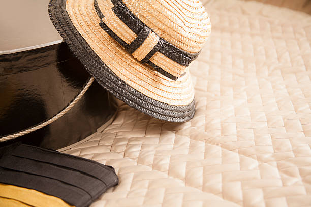 Woman's straw hat in tan and black colors, woman's handbag, and a black, round hat box lie on top of a bed in a bedroom setting.  No people.  Copyspace.  Retail, shopping, consumerism, tourism, travel, vacation concepts.