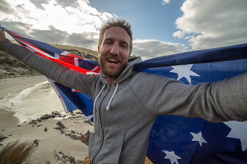 Cheerful young man on a beach in Kangaroo Island takes a selfie portrait holding an Australian's flag.