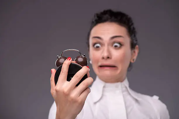 Close-up portrait of pretty girl with surprised face holding an alarm clock in her hand and looking at it with a fright, with copy place isolated on grey background, concept of time management