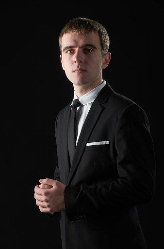 Young man in black suit and white shirt with hands in his pockets on black background
