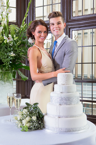 A happy young couple smile at the camera while standing by their wedding cake.