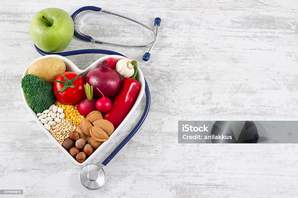 Healthy Foods in Heart Shaped with Stethoscope Heart health, and cholesterol diet concept.  Healthy foods in heart shaped bowl with stethoscope and green apple on white vintage wooden table. Healthy Eating Stock Photo