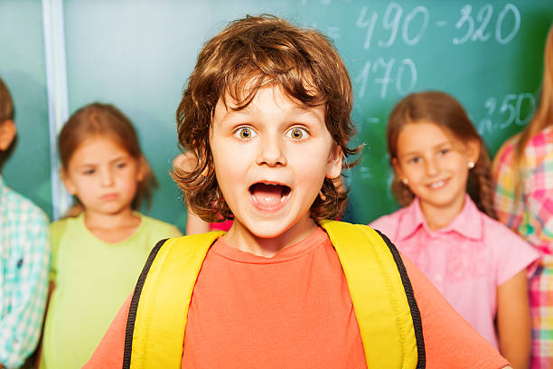 Portrait of excited boy with bag near chalkboard Portrait of excited boy with yellow bag near chalkboard and other students during mathematics class blackboard child shock screaming stock pictures, royalty-free photos & images