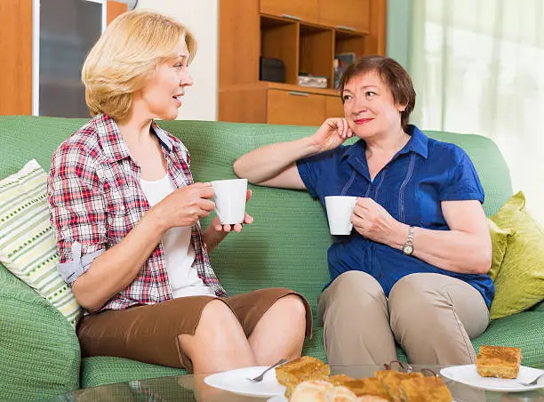 Two positive mature friends drinking tea and gossiping at home