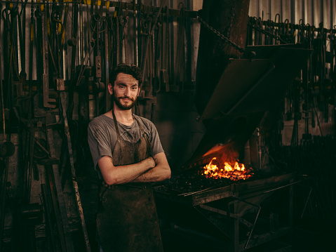 Portrait of a fulfilled young adult craftsman standing in his blacksmith factory