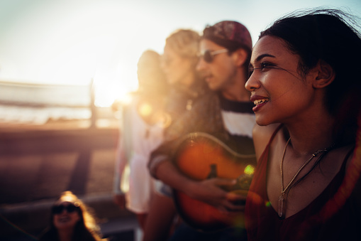 Hipster friends relaxing at the beach with an acoustic guitar during a summer road trip at sunset with sun flare