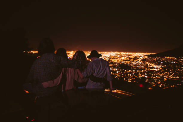 Back view of a group of friends hugging sitting on the bonnet of a car and watching night city lights from viewpoint